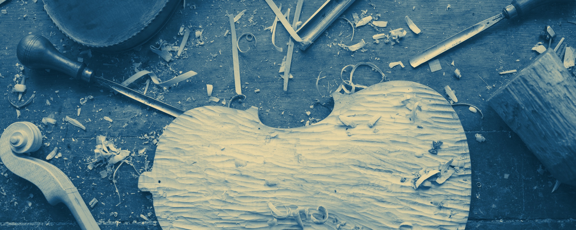 A luthier's bench seen from above: a violin back part way through
            being carved, gouges and chisels laid beside it, and a finished
            instrument at the edge of the bench.